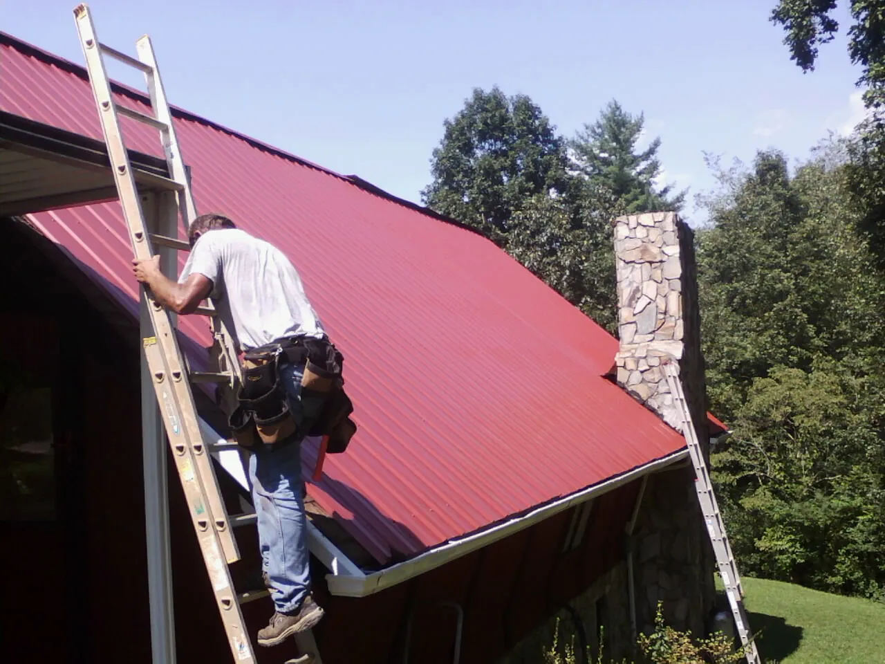 Roofer on a ladder beside a red metal roof and stone chimney — Soffit & Fascia Repair by Roofing Brothers in Hixson